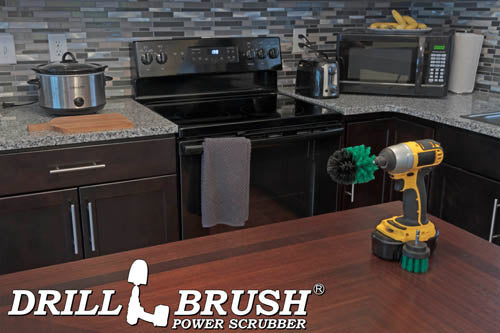 The Drillbrush 2M Medium Green Kitchen Brush Kit in a cordless drill sitting on a dinner table in front of kitchen counters, a stove, a toaster, and a microwave.
