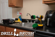 The Drillbrush 4M Medium Green Kitchen Brush Kit in a cordless drill sitting on a countertop in front of a sink, coffeemaker, and dishes.