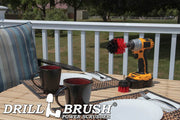The Drillbrush 2M Red Stiff Outdoor & Patio Brush Kit in a cordless drill sitting on a deck table set with forks, mugs, and paper plates in from of a grassy backyard.