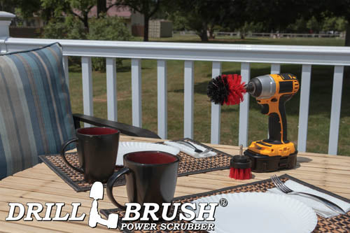 The Drillbrush 2M Red Stiff Outdoor & Patio Brush Kit in a cordless drill sitting on a deck table set with forks, mugs, and paper plates in from of a grassy backyard.