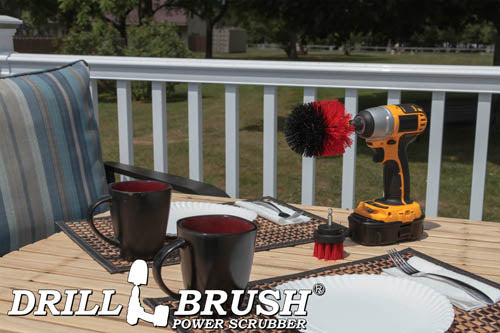 The Drillbrush 2O Red Stiff Outdoor & Patio Brush Kit in a cordless drill sitting on a deck table set with forks, mugs, and paper plates in from of a grassy backyard.