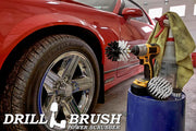 The Drillbrush 42O Soft White Home & Auto Brush Kit in a cordless drill sitting on a bucket next to a spray bottle and a rag in front of a red sports car.