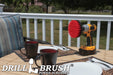 Drillbrush 5 inch Stiff Red Outdoor & Patio Brush on a deck table next to paper plates, silverware, and mugs in front of a backyard.