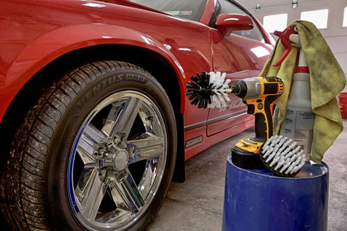 The Drillbrush 4O Soft White Home & Auto Brush Kit in a cordless drill sitting on a bucket next to a spray bottle and a rag in front of a red sports car.