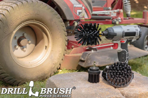 The Drillbrush 4OS-2L Ultra Stiff Black Grill & Industrial Brush Kit in a cordless drill sitting on bricks in front of a ride-on lawnmower.