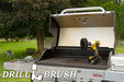 The Drillbrush 5O Ultra Stiff Black Grill & Industrial Brush Kit in a cordless drill sitting on an open grill in front of a grassy backyard.