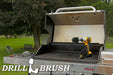 The Drillbrush MO Ultra Stiff Black Grill & Industrial Brush Kit in a cordless drill sitting on an open grill in front of a grassy backyard.