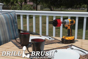 Drillbrush Mini Stiff Red Outdoor & Patio Brush on a deck table next to paper plates, silverware, and mugs in front of a backyard.
