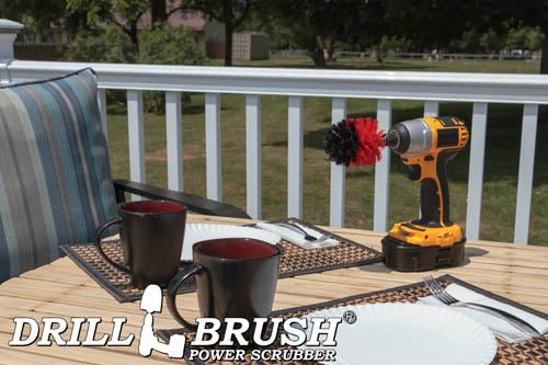 Drillbrush Mini Stiff Red Outdoor & Patio Brush on a deck table next to paper plates, silverware, and mugs in front of a backyard.
