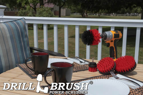 The Drillbrush 542O Red Stiff Outdoor & Patio Brush Kit in a cordless drill sitting on a deck table set with forks, mugs, and paper plates in from of a grassy backyard.