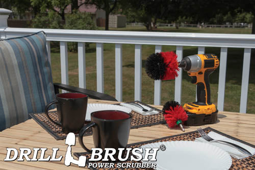 The Drillbrush MO Red Stiff Outdoor & Patio Brush Kit in a cordless drill sitting on a deck table set with forks, mugs, and paper plates in from of a grassy backyard.