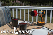 Drillbrush Original Stiff Red Outdoor & Patio Brush on a deck table next to paper plates, silverware, and mugs in front of a backyard.