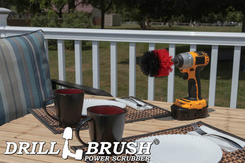 Drillbrush Original Stiff Red Outdoor & Patio Brush on a deck table next to paper plates, silverware, and mugs in front of a backyard.