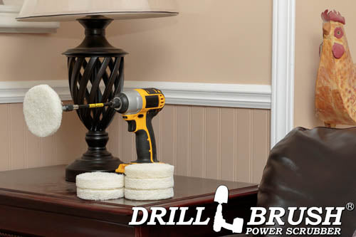 The Drillbrush 6 Soft White 4 inch Scrubbing Pads and 5 inch extension Kit, with one pad on a 3 inch backer attached to an extension in a cordless drill on a wooden end table next to a leather sofa in front of a lamp.