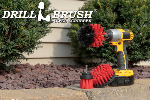 The Drillbrush 4OS-2L Red Stiff Outdoor & Patio Brush Kit in a cordless drill sitting on a brick planter in front of a rock garden and shrubs.