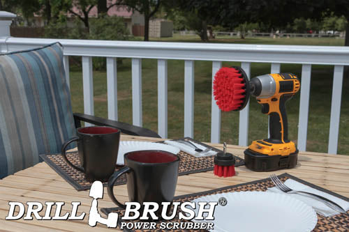 The Drillbrush 42 Red Stiff Outdoor & Patio Brush Kit in a cordless drill sitting on a deck table set with forks, mugs, and paper plates in from of a grassy backyard.