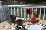 The Drillbrush 4O Red Stiff Outdoor & Patio Brush Kit in a cordless drill sitting on a deck table set with forks, mugs, and paper plates in from of a grassy backyard.