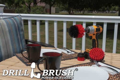 The Drillbrush 42O Red Stiff Outdoor & Patio Brush Kit in a cordless drill sitting on a deck table set with forks, mugs, and paper plates in from of a grassy backyard.