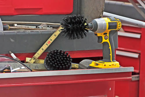 The Drillbrush 4O Ultra Stiff Black Grill & Industrial Brush Kit in a cordless drill sitting on an open shelf of a tool box in a garage.